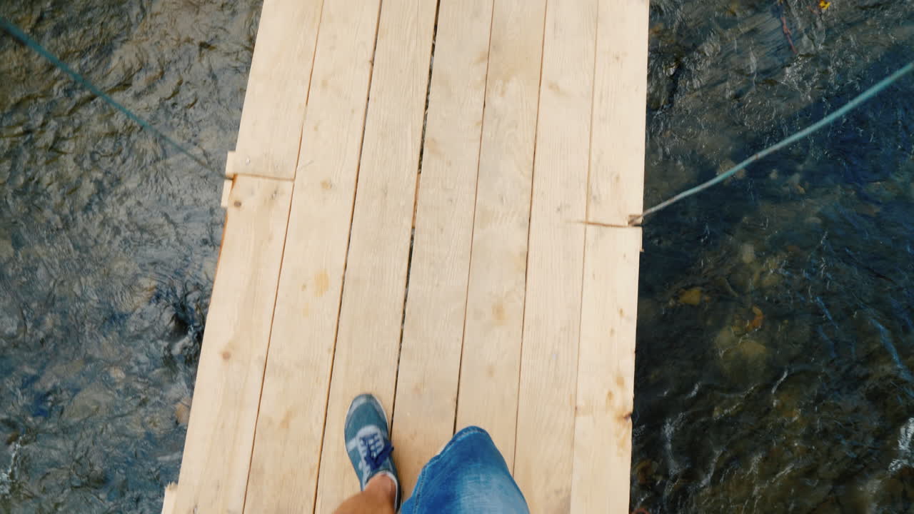 en mand går langs en smal, vaklende bro over floden. i billedet kan man kun se benene.