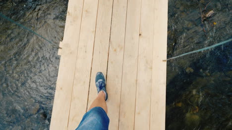 A-Man-Walks-Along-A-Narrow-Wobbly-Bridge-Across-The-River-In-The-Frame-One-Can-See-Only-The-Legs-Pov