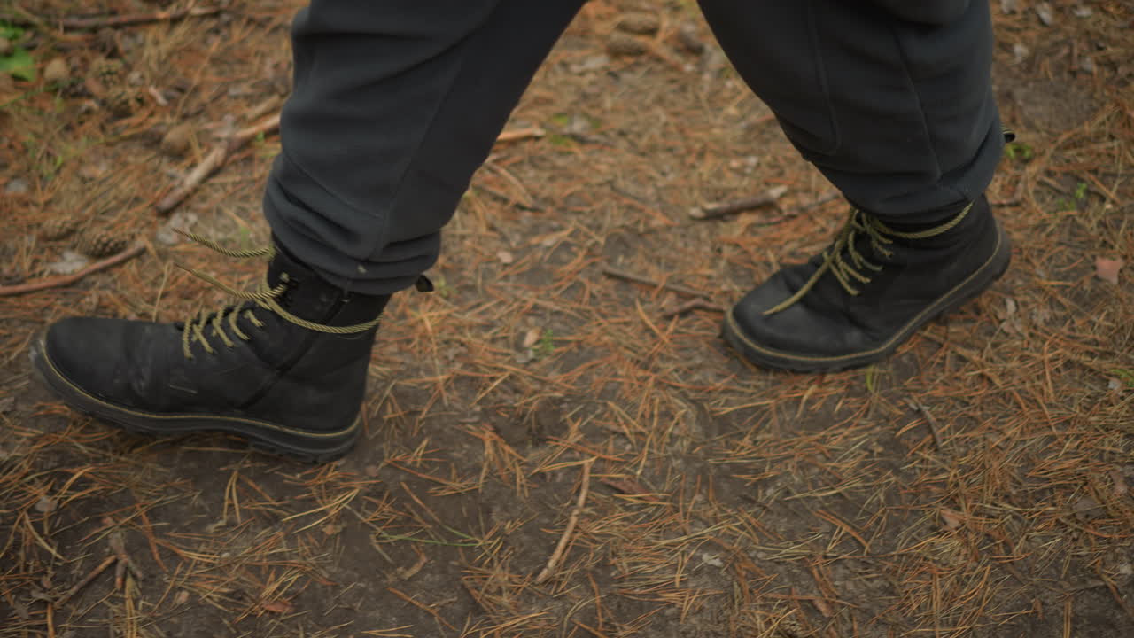 closeup sepatu hiking menginjak jarum pinus, langkah hati-hati perlahan di jalan tanah, sepatu bertali gelap dan celana abu-abu, bunyi berderit halus dan tanah bertekstur, penjelajah solo merasakan suasana hutan yang tenang