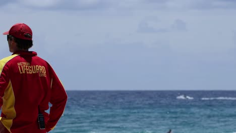 a lifeguard in uniform stands watchfully by the ocean, ensuring safety under a partly cloudy sky.
