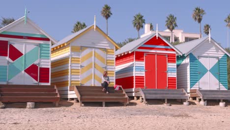 Eine-Dame,-Die-Auf-Den-Stufen-Einer-Der-Ikonischen,-Bunt-Bemalten-Strandhütten-Am-Strand-Von-Brighton,-Victoria,-Sitzt