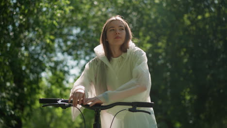 young woman in translucent raincoat removes hood and leans on bicycle handlebar looking thoughtful under dappled sunlight filtering through green leaves