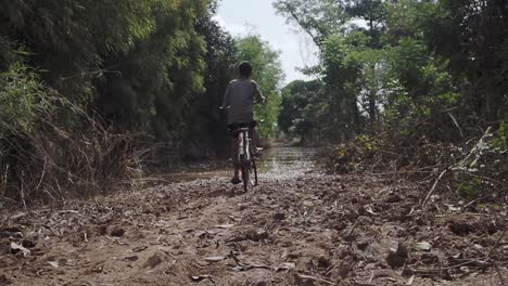 young boy trying to cycle with old bike through flooded rural road after heavy rain in cambodia