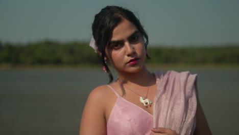 young woman in pink dress posing by a serene lake, daylight, candid expression