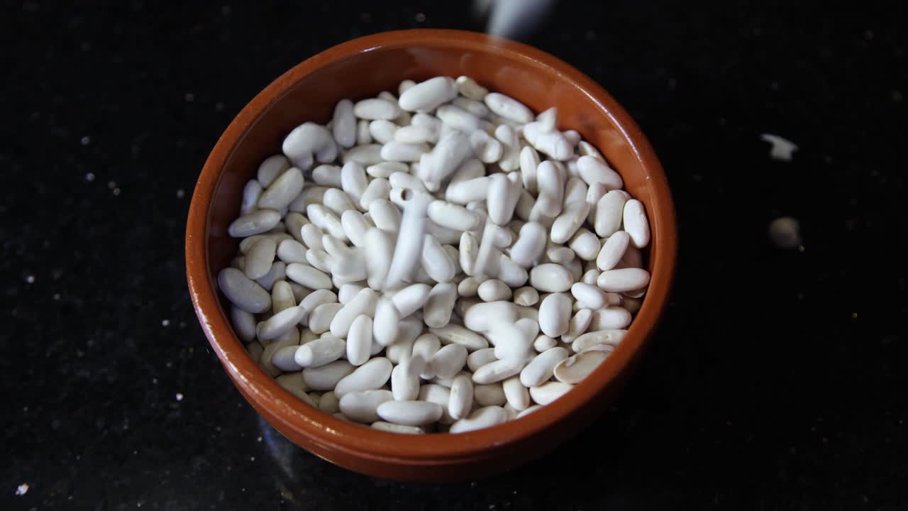 Close up of white beans tumbling into a rustic terracotta bowl against a dark speckled background, hinting at culinary preparation and fresh ingredients