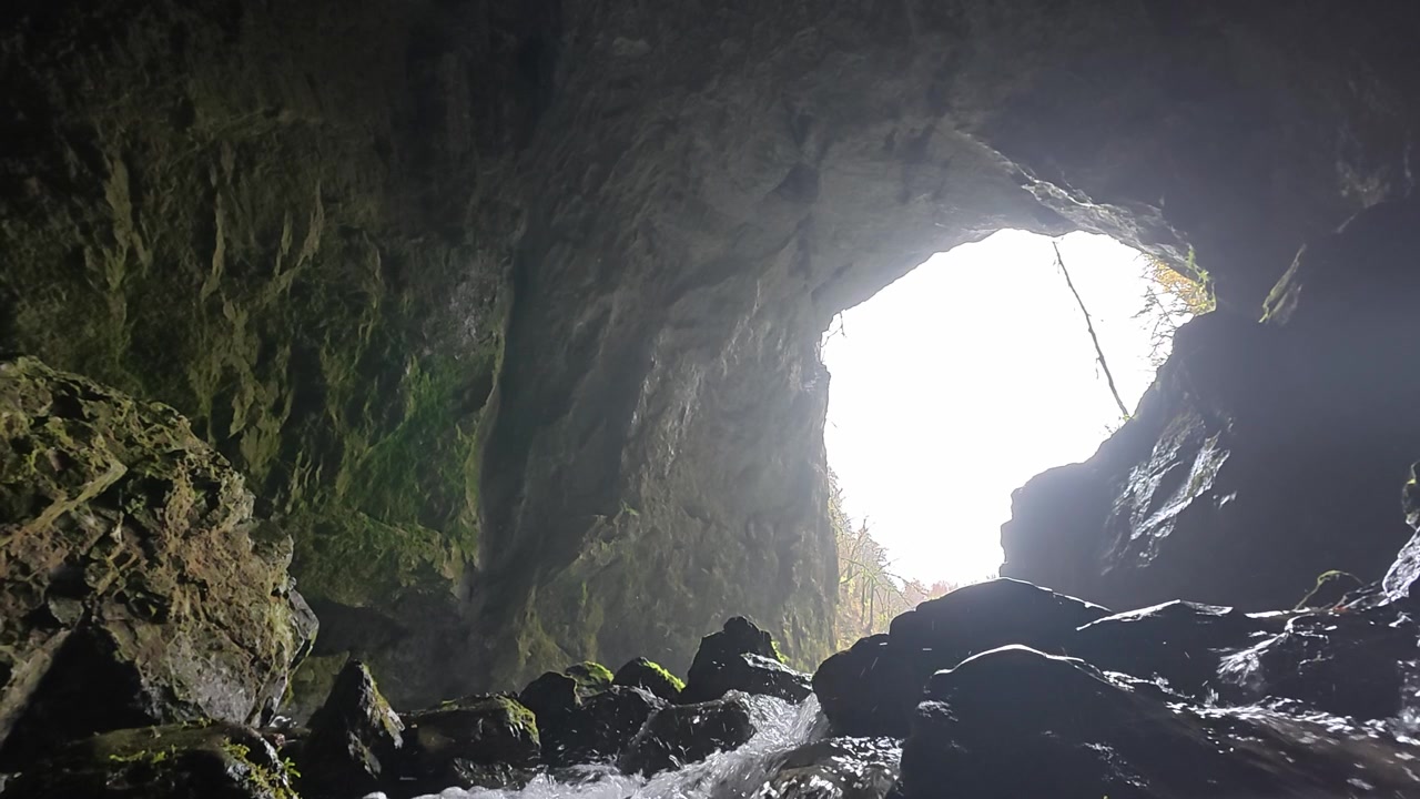 Bright sunlight illuminating the entrance of a dark cave while a river flows over rocks. The stream emerges from the underground cavern