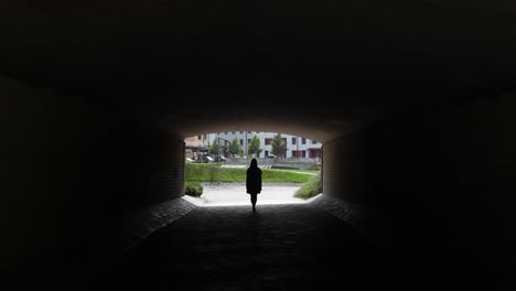 silhouette woman walking in dark tunnel walking out from underpass to bright street in latvia, back view