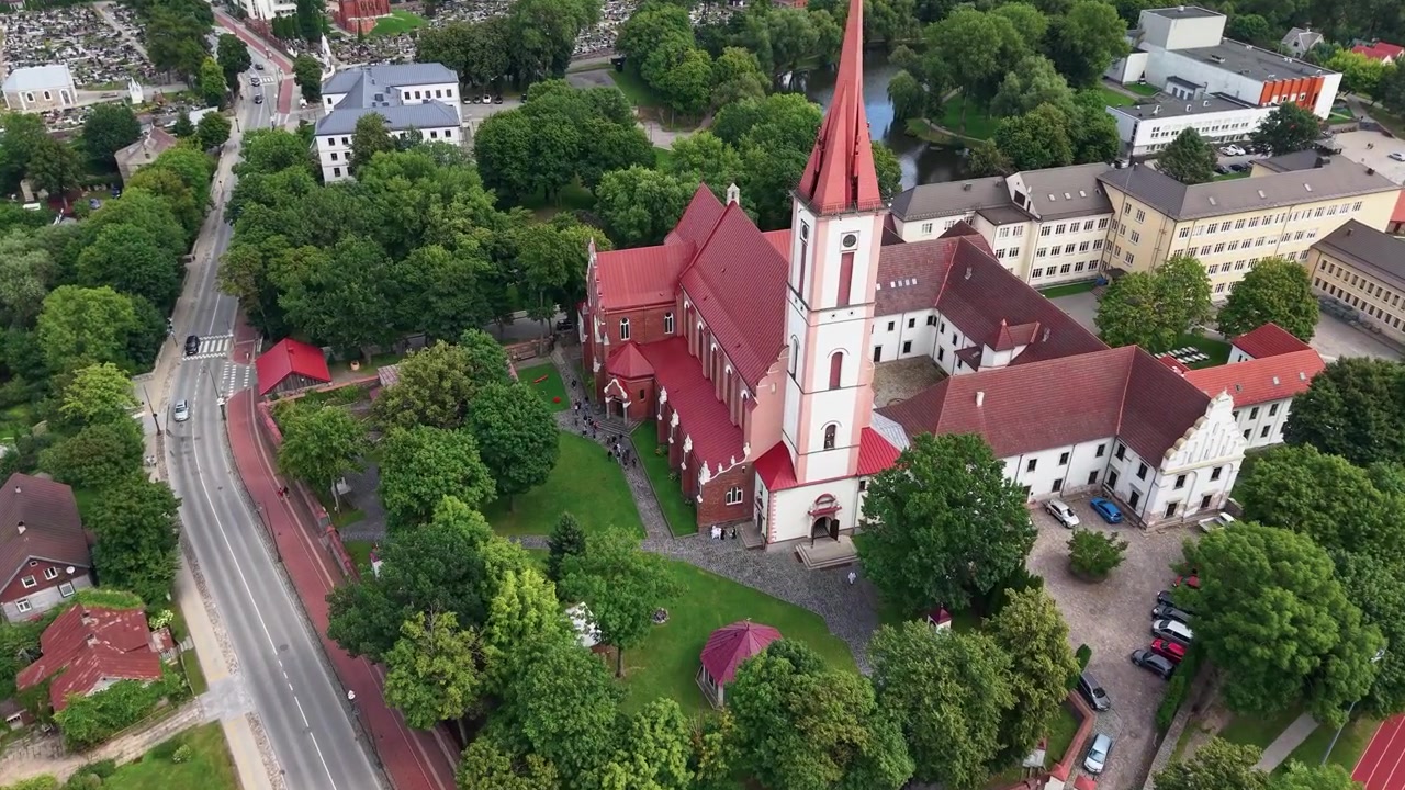 Fantastiska flygbilder visar Kyrkan för Herrens uppenbarelse till Jungfru Maria i Kretinga, Litauen, omgiven av frodig grönska, röda tak och ett livfullt stadslandskap i mjukt dagsljus.