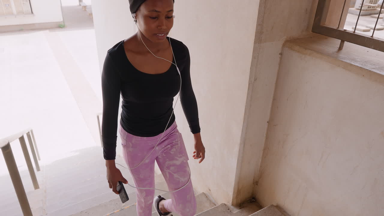 black woman walking up stairs, ascending concrete stairwell with purposeful stride, carrying keys, wearing black top and purple leggings, morning sunlight casting texture on walls, closeup