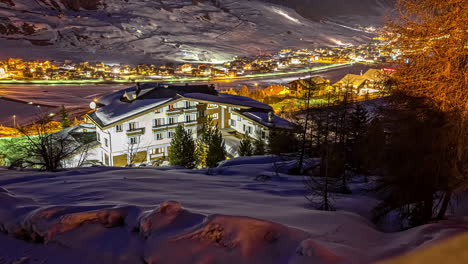 Zeitrafferaufnahme-Der-Schneebedeckten-Alpen-Im-Tal-Mit-Beleuchtetem-Dorf-Am-Abend-–-Fahrende-Pistenraupen-Auf-Der-Piste