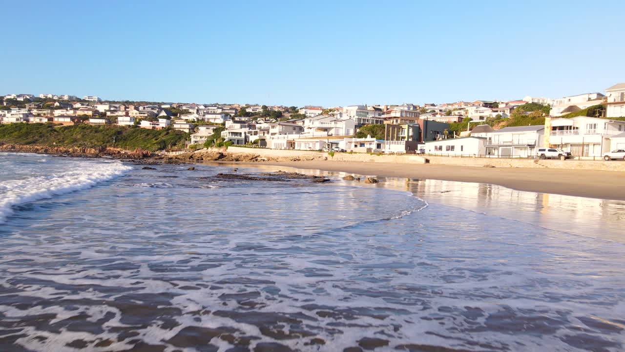 Low sunrise aerial pullback over waves running out onto Vleesbaai beach village