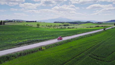 Toma-Aérea-De-Un-Auto-Rojo-Conduciendo-Por-Un-Paisaje-Verde-De-Toscana