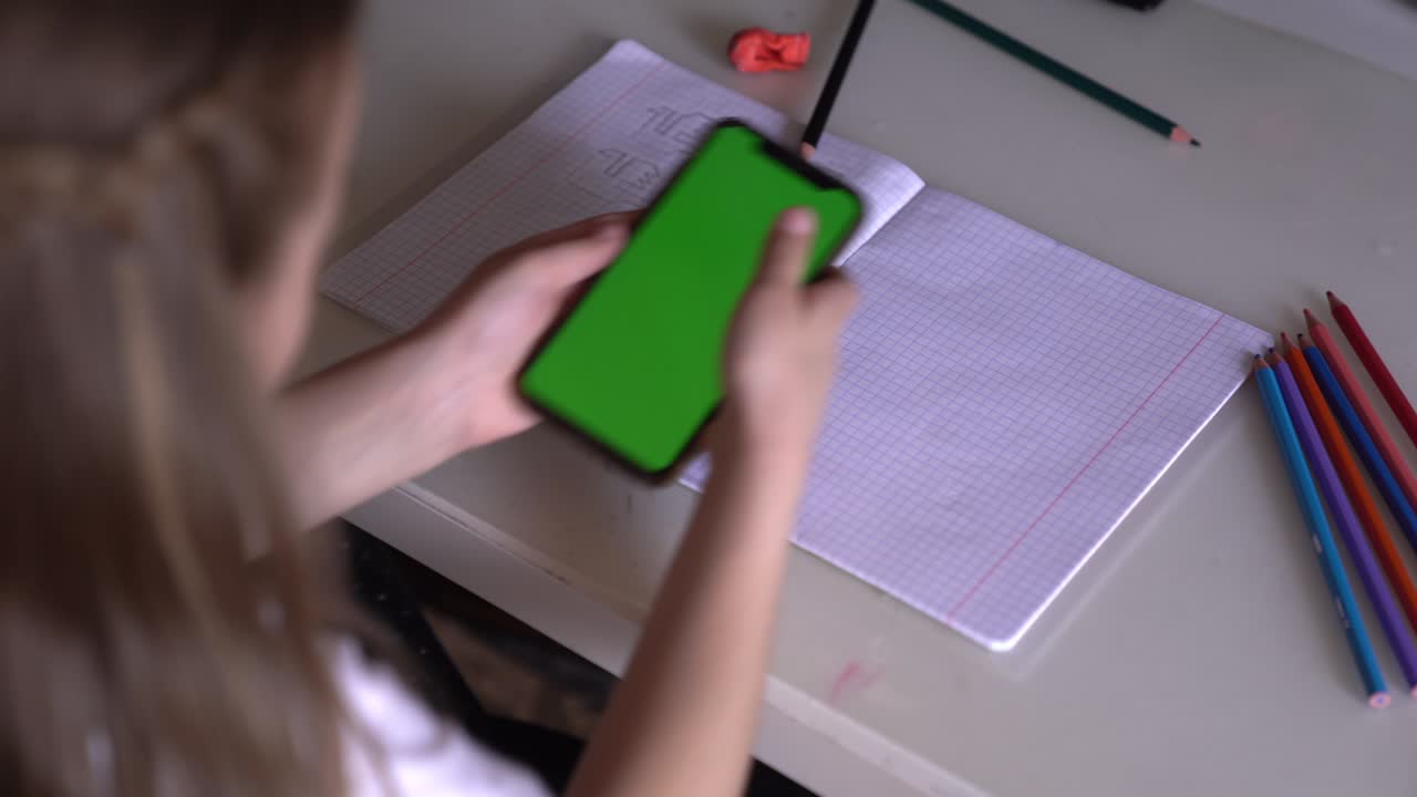 una niña está sentada en una mesa con el teléfono en las manos, mira el croma