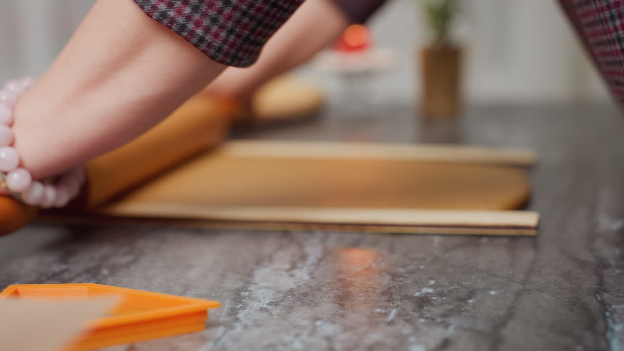 Close-up of woman hands rolling brown dough with a wooden rolling pin, left wrist adorned with a pink beaded bracelet, slight blur in background featuring candle and baking tools