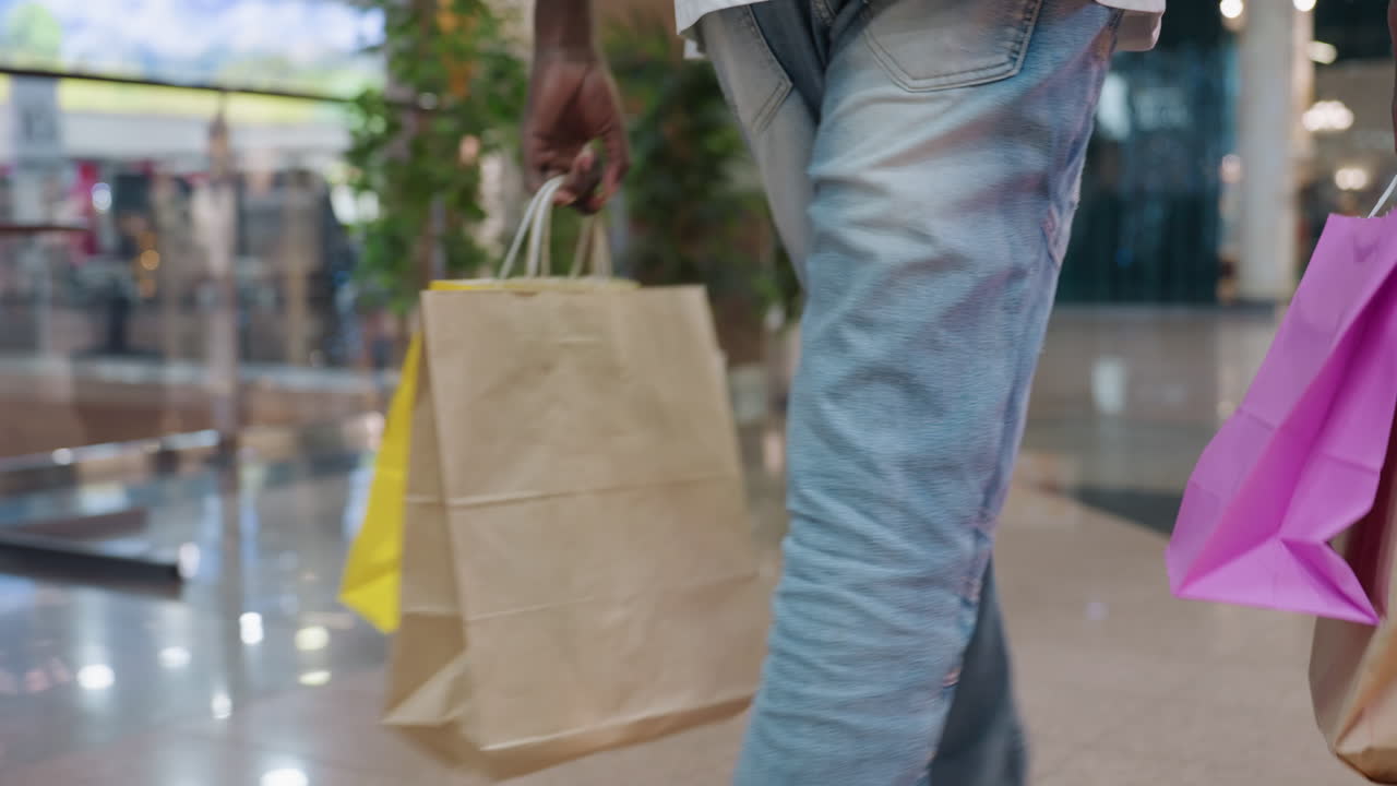 Close up of man holding paper shopping bags in both hands while walking through brightly lit indoor shopping center with reflections on polished floor creating consumer lifestyle atmosphere
