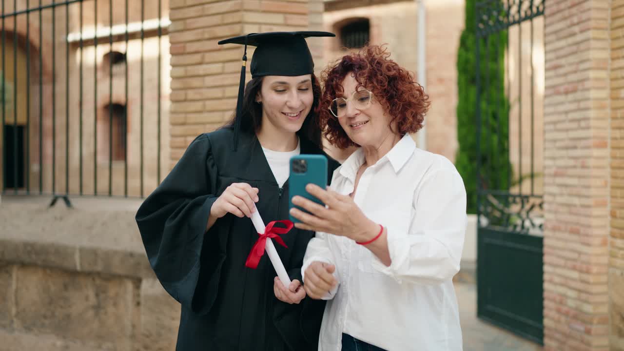 Two women mother and daughter holding graduate diploma make selfie by smartphone at campus university