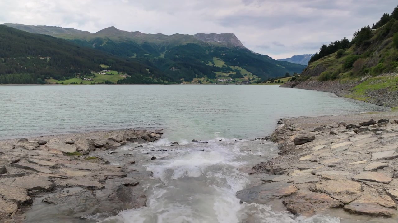 Tranquillo bacino idrico montano del Lago di Resia in Alto Adige con sponda rocciosa e flusso d'acqua delicato sotto un cielo nuvoloso, circondato da verdi colline e vette distanti.