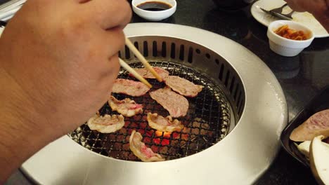 grilled thinly sliced beef turned with chopsticks and another pair of chopsticks held by a woman added sausages on the grill