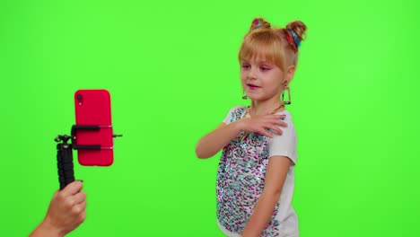 Cheerful-funny-little-teen-girl-kid-dancing-at-camera-filming-video-using-phone-on-chroma-key-wall