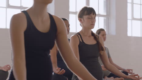 joven mujer caucásica embarazada en clase de yoga practicando postura de loto disfrutando de la práctica de meditación en grupo relajándose en el gimnasio al amanecer.