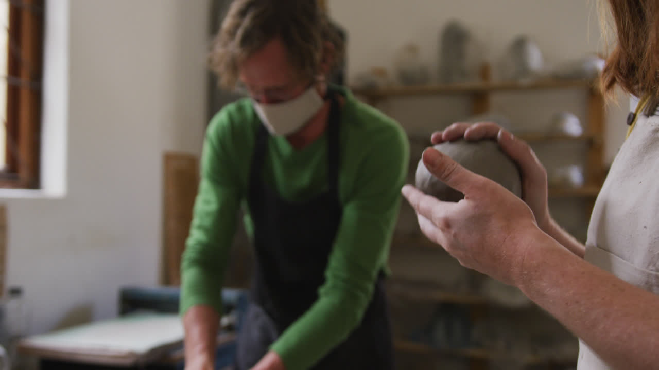 hombre y mujer alfareros con máscara facial y delantal amassando la arcilla en el estudio de cerámica