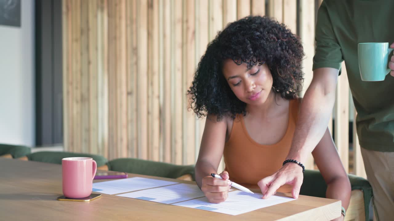 Diverse couple at wooden dining table, man entering with green mug, guiding woman writing paperwork