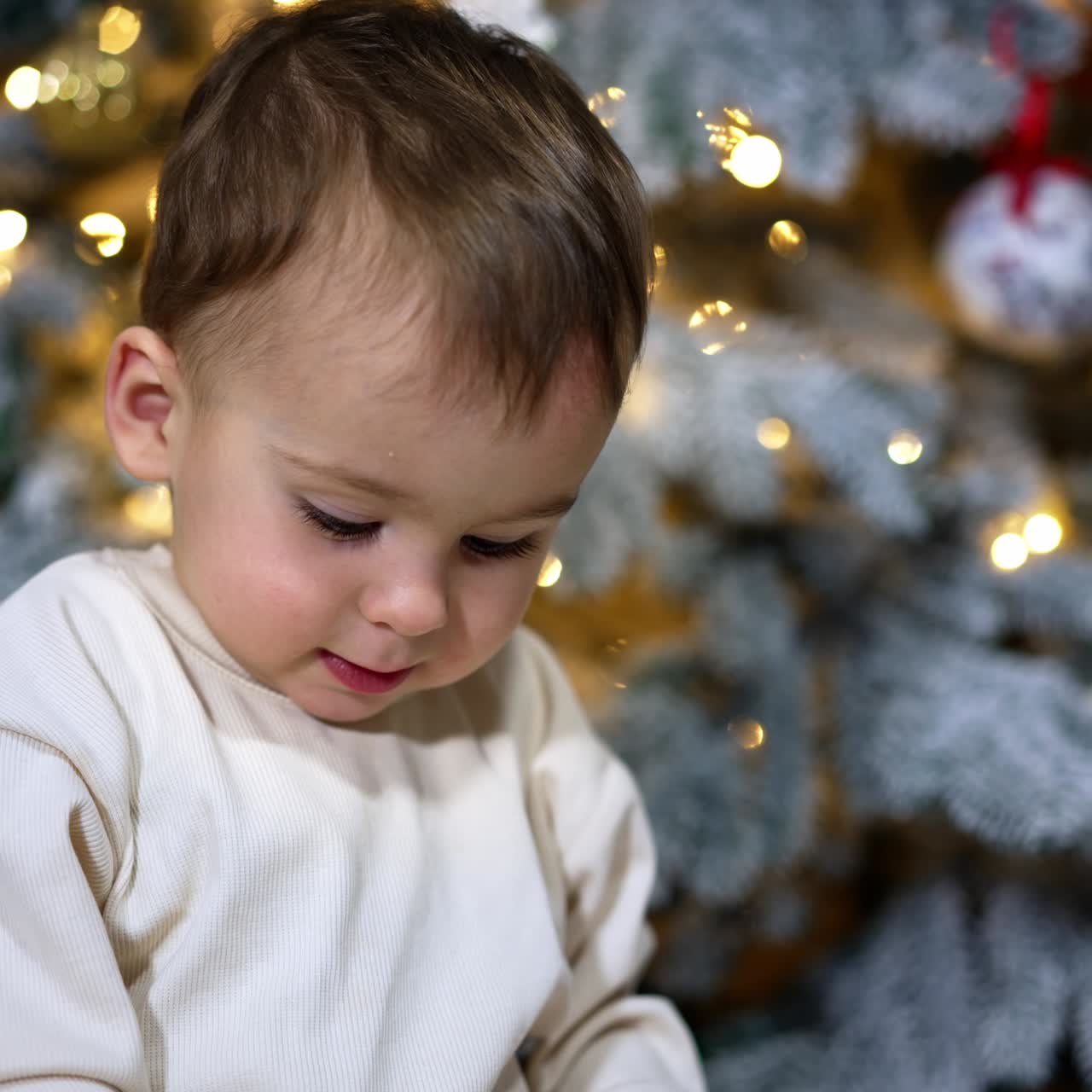 Beautiful angelic kid in white clothes sits near the nicely decorated Christmas tree. Lovely child plays with a toy and waves his hands happily