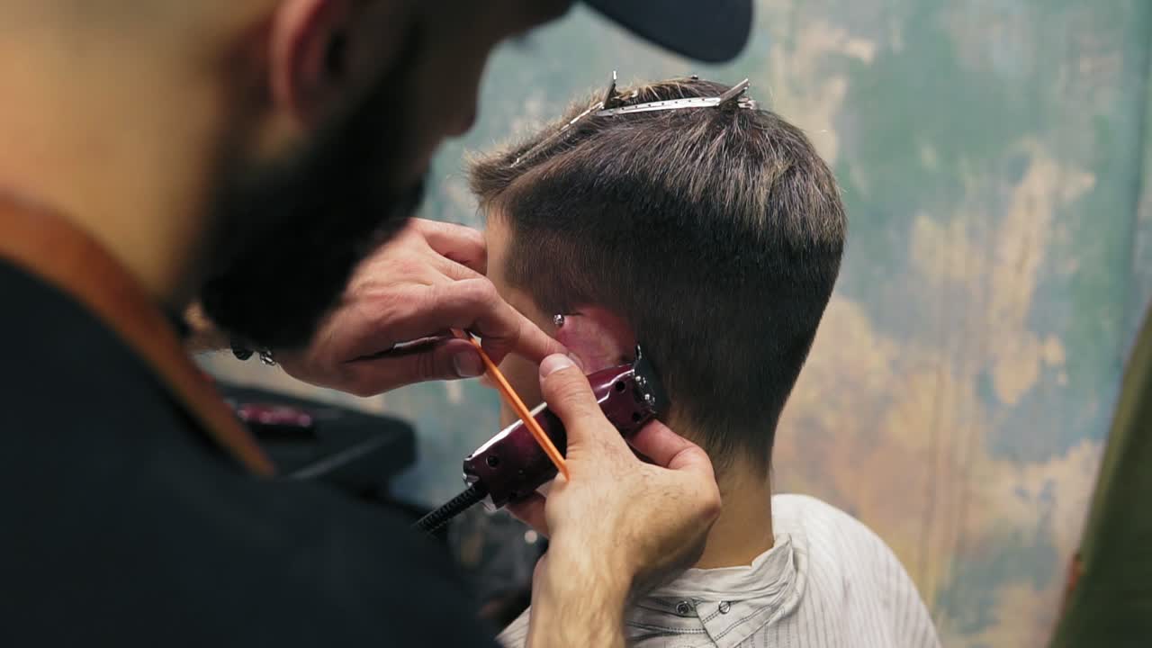 Bearded barber creating a straight line using electric trimmer behind a man's ear. Young handsome caucasian man getting a