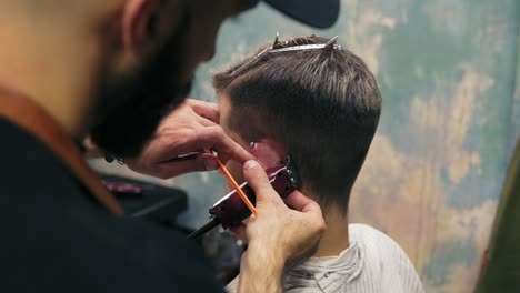 Bearded-barber-creating-a-straight-line-using-electric-trimmer-behind-a-man's-ear.-Young-handsome-caucasian-man-getting-a