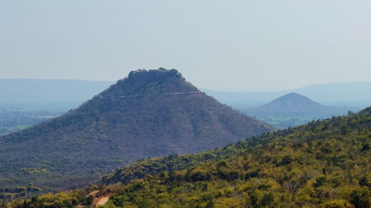 Veduta panoramica lontana della collina di Trikuta con il tempio di Maa Sharda sulla cima, circondata da colline boscose e un paesaggio rurale a strati sotto un cielo azzurro velato