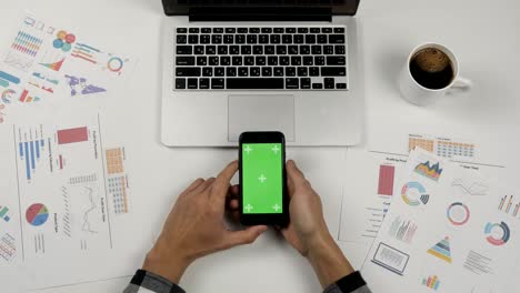 hombre de negocios sosteniendo un teléfono inteligente con pantalla verde en el fondo de la mesa de oficina blanca. manos masculinas desplazando páginas.