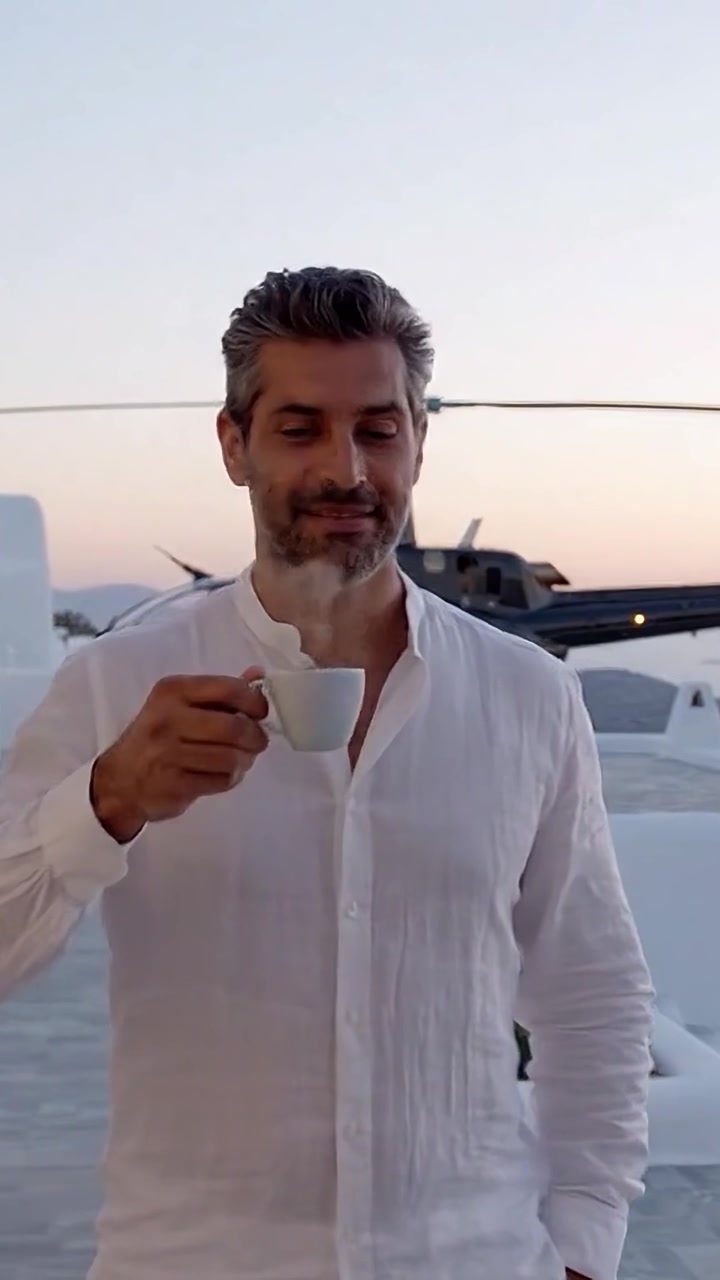 Uomo in abito bianco formale si gode un caffè mentre è in piedi vicino a un elicottero su una terrazza moderna con un tramonto panoramico sullo sfondo.