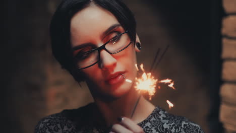 Portrait-Of-Young-Goth-Woman-With-Short-Black-Hair-Wearing-Glasses-He-Looks-At-A-Burning-Sparkler-Sl