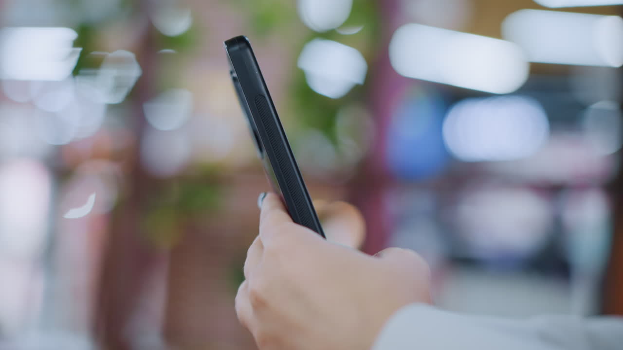 휴대폰을 조종하는 사람의 가까운 손으로 찍은 사진, 장치 쪽에 초점을 맞추고 부드러운 흐릿한 배경, 현대적이고 유행적인 설정에서 스마트폰 사용과 라이프 스타일을 강조합니다.