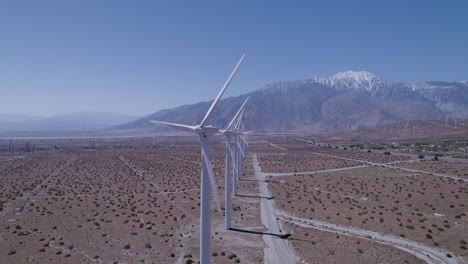 un drone lento revela una línea de molinos de viento en funcionamiento en el desierto, con montañas cubiertas de nieve en el fondo