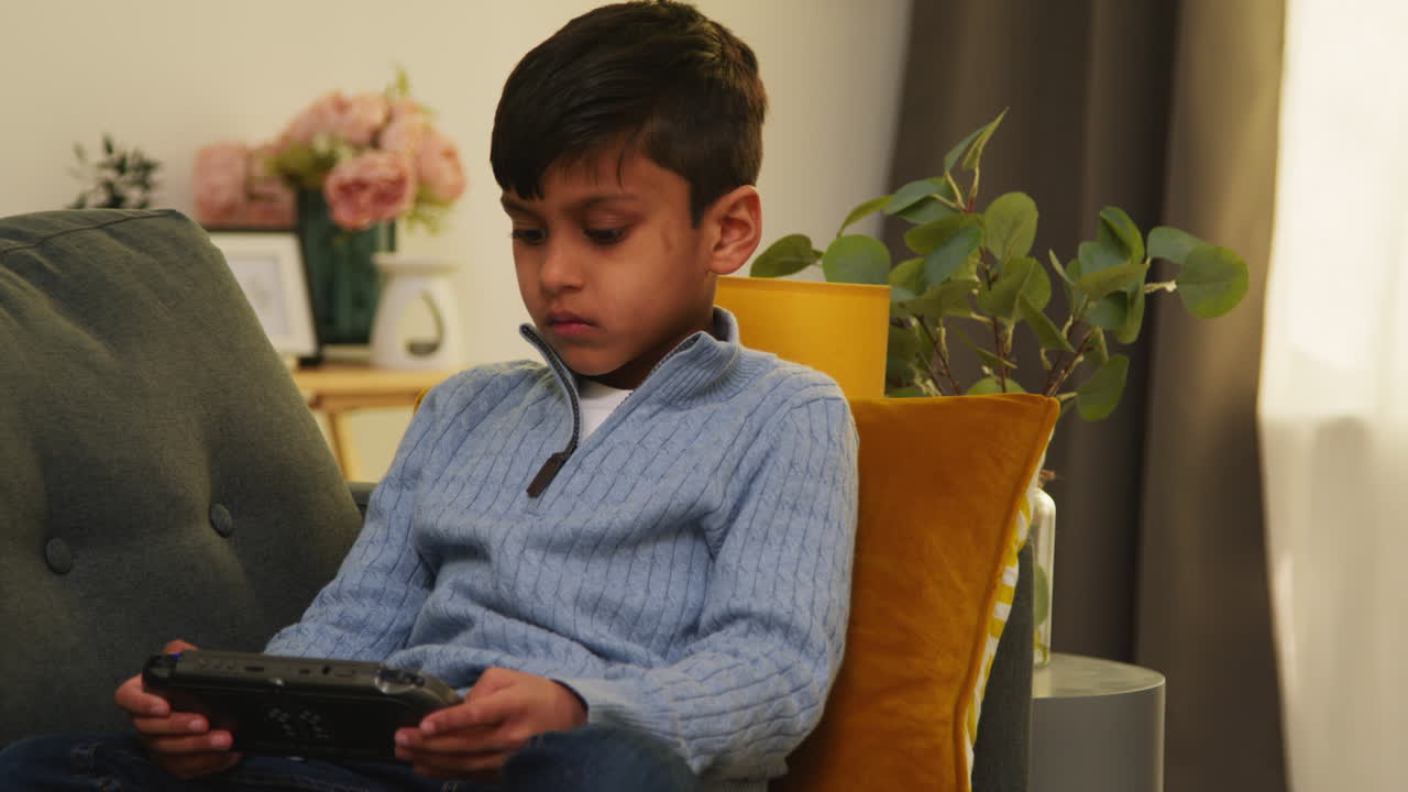 Young Boy Sitting On Sofa At Home Playing Game Or Streaming Onto Handheld Gaming Device 1