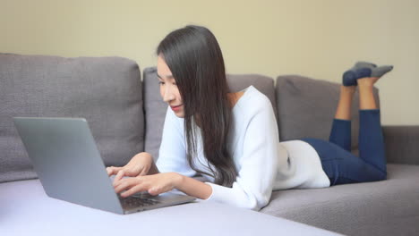 young asian woman typing on her laptop lying down on sofa