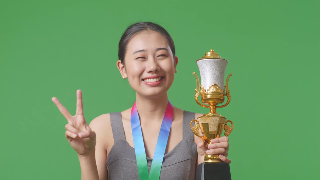 금메달과 트로피를 들고 있는 아시아 여성이 평화의 몸짓을 하고 카메라를 향해 미소 짓는 모습이 스튜디오의 초록색 스크린 배경에서 첫 번째 우승자입니다.