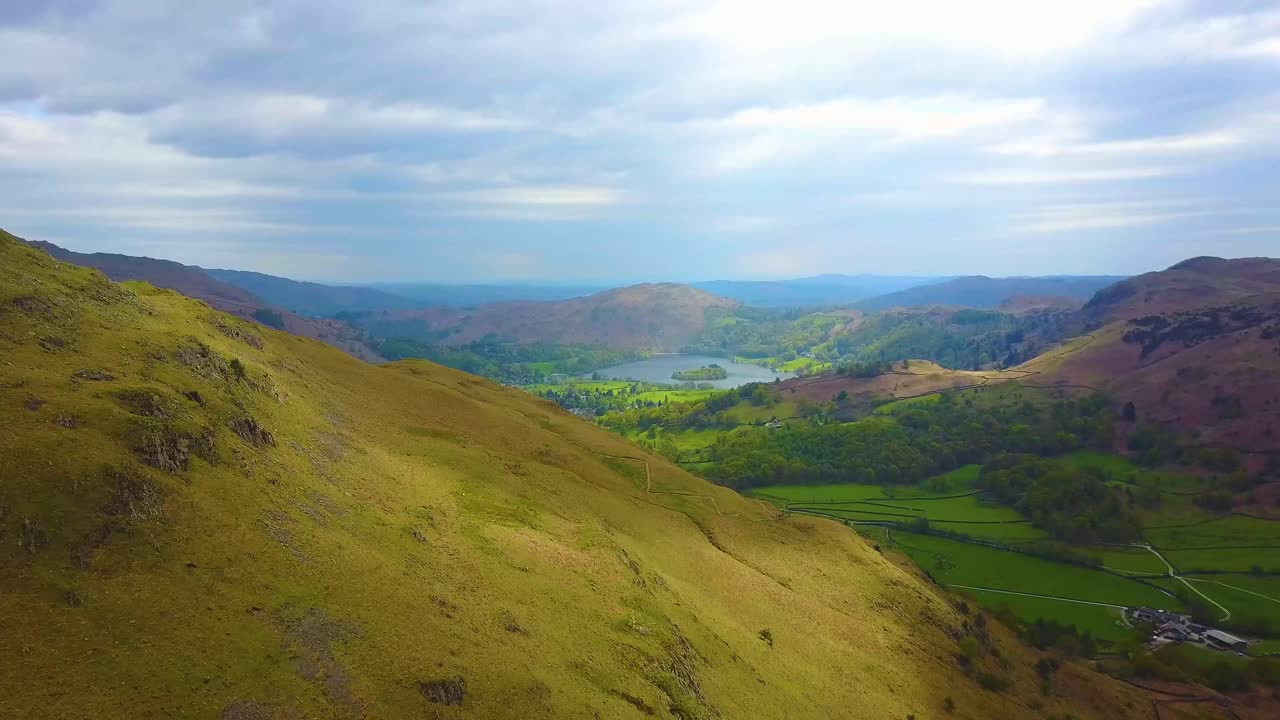 An aerial view from a drone flying over rolling green hills towards a lake in the distance.  The sunshine is casting shadows from the clouds which appear to be seen racing across ground.