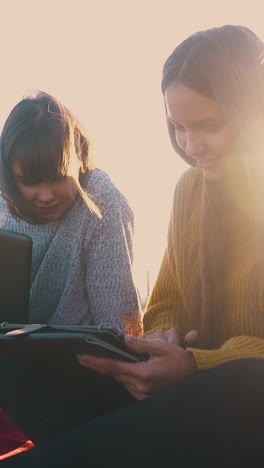 smiling women with tablet and laptop sit in comfortable campsite with tent against back sunset light in autumn evening