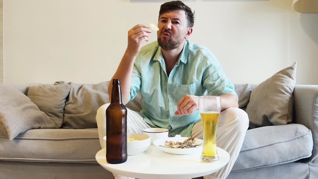 Man relaxes on a couch, eating snacks and drinking beer