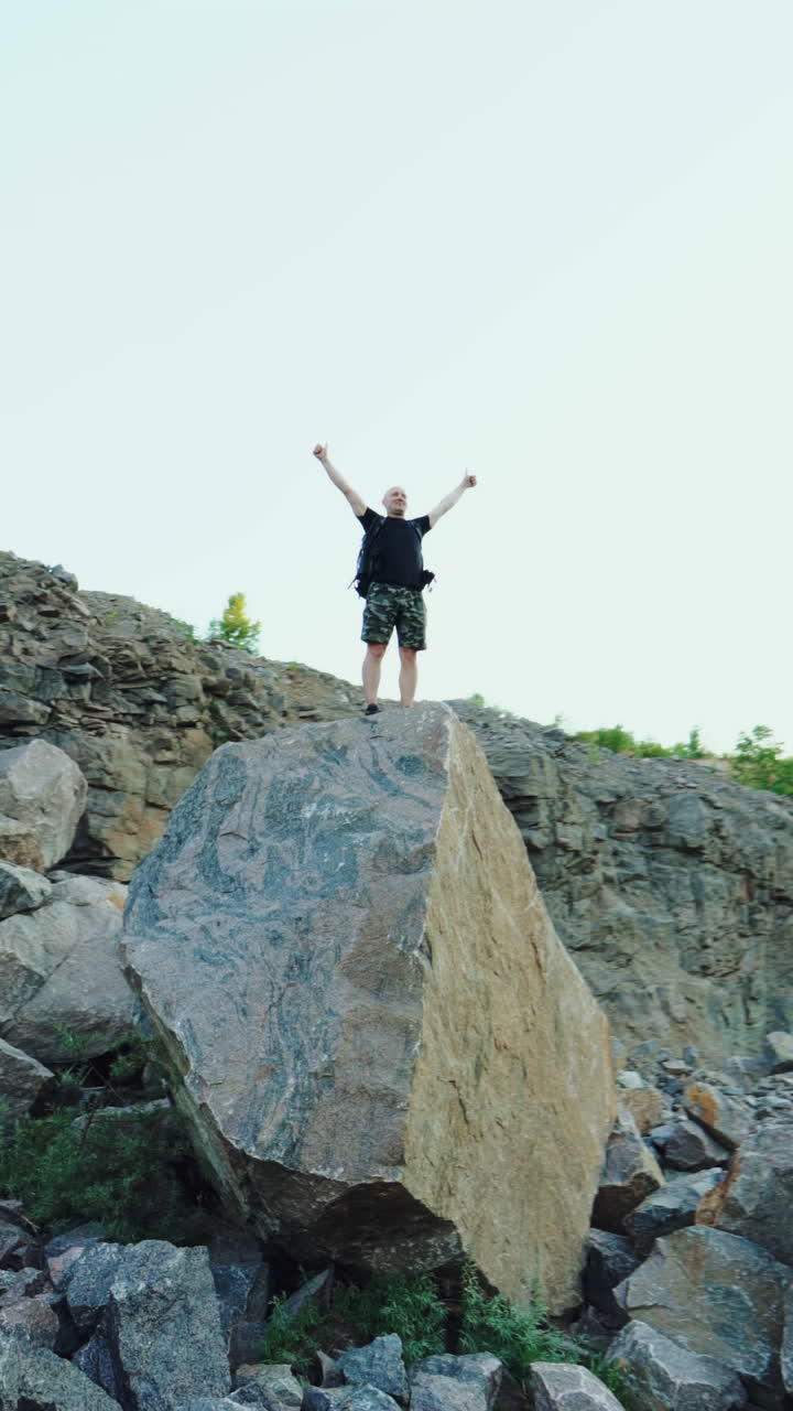 Tourist man standing on a big rock and raises his hands up on the stones background under the clear sky. Happy young hiker man tourist between the rocks outdoors. Vertical video