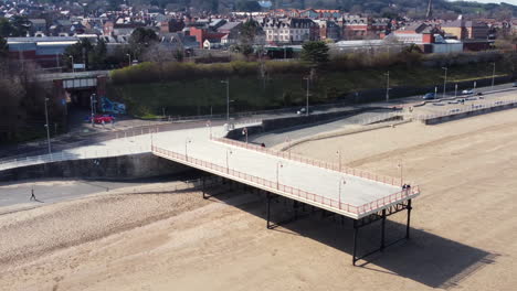 An-aerial-view-of-Colwyn-Bay-seafront-and-small-fishing-jetty-on-a-sunny-day,-flying-left-to-right-around-the-jetty-while-zooming-in,-Denbighshire,-North-Wales,-UK