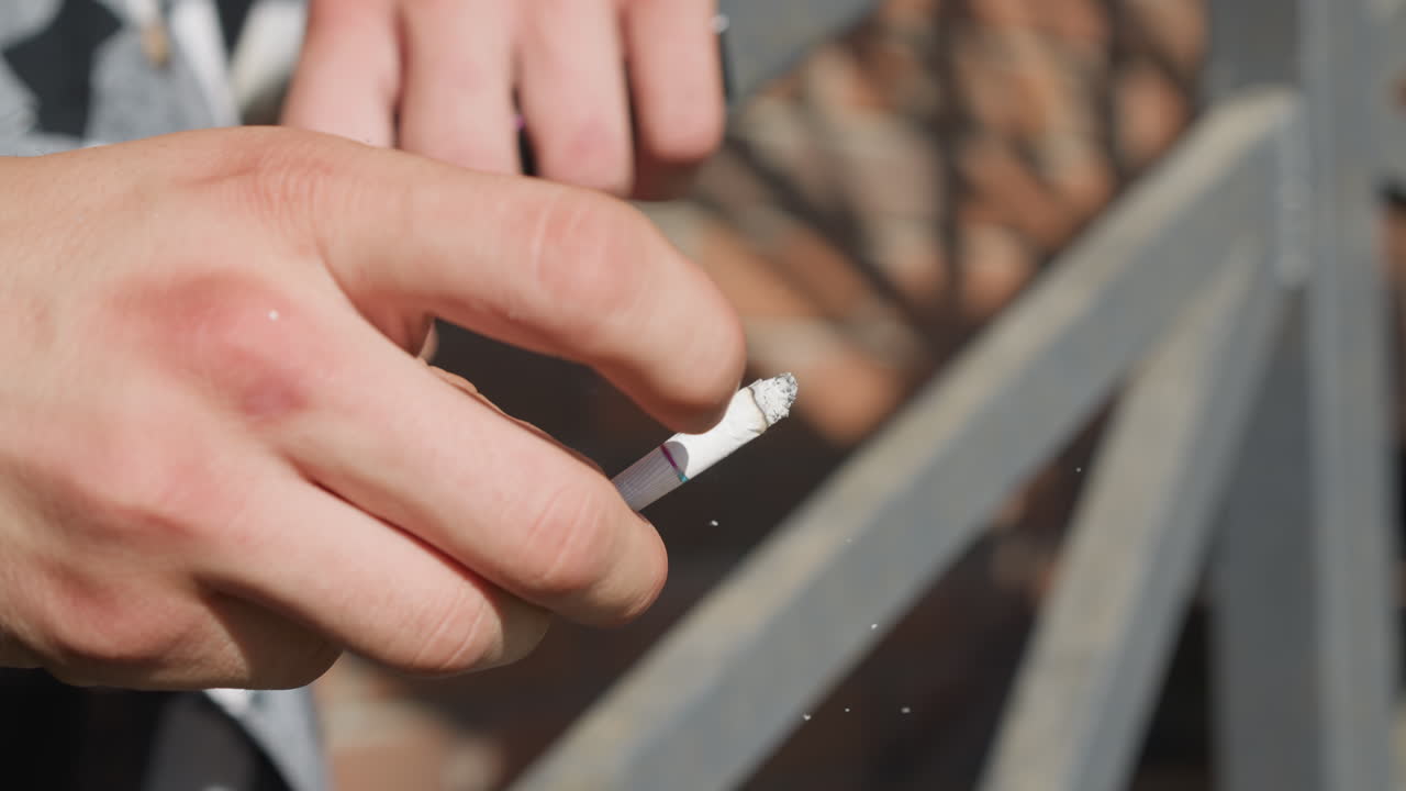 Close up of smoker hand flicking cigarette as ash breaks off midair while leaning on iron railing in outdoor sunlit area with soft focus background and diffused light catching falling ash particles