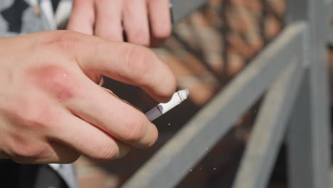 Close up of smoker hand flicking cigarette as ash breaks off midair while leaning on iron railing in outdoor sunlit area with soft focus background and diffused light catching falling ash particles