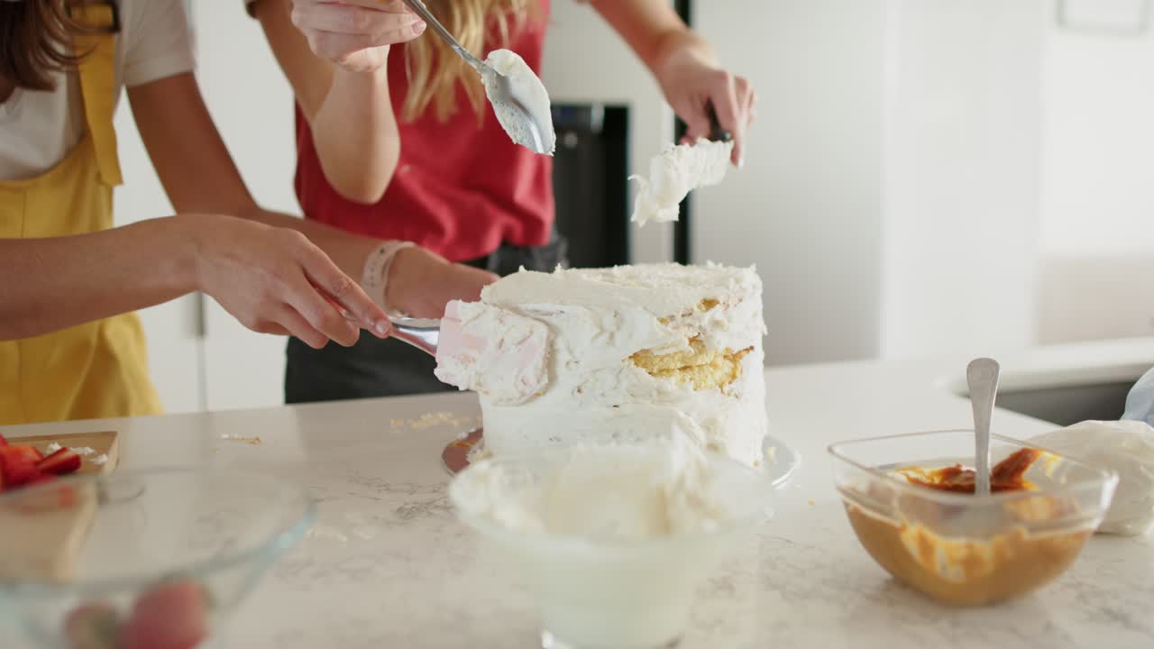 Female friends scooping frosting and smoothing it onto three-layer cake in kitchen for decorating
