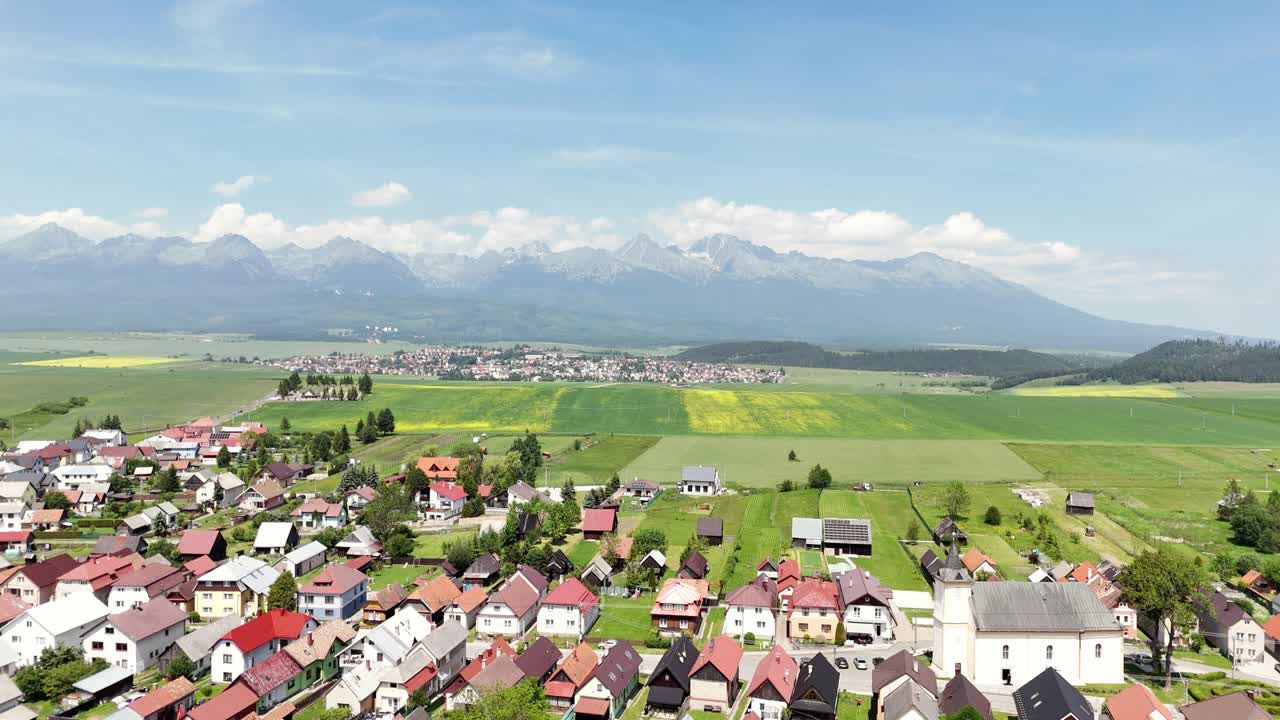 Aerial view of a peaceful rural landscape with scattered houses, a small church, and wide green fields stretching toward the majestic High Tatras mountains under a bright summer sky
