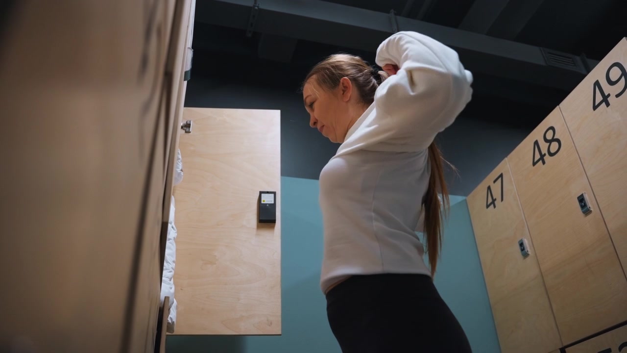 Badminton player in locker room slipping into sweatshirt and zipping collar, tucking hair, checking locker numbers, soft warm lighting, postpractice cooldown ritual, recreational player breathing out