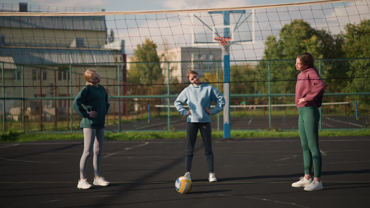 야외 배구장 에 서 있는 세 명 의 여자 들 은 머리 를 왼쪽 과 오른쪽 으로 구부려 운동 하고 있다. 배경 에 스포츠 경기장 이 있고, 앞 에 배구장 이 있다.