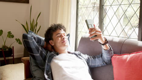 biracial man lying on couch and holding smartphone at home, slow motion